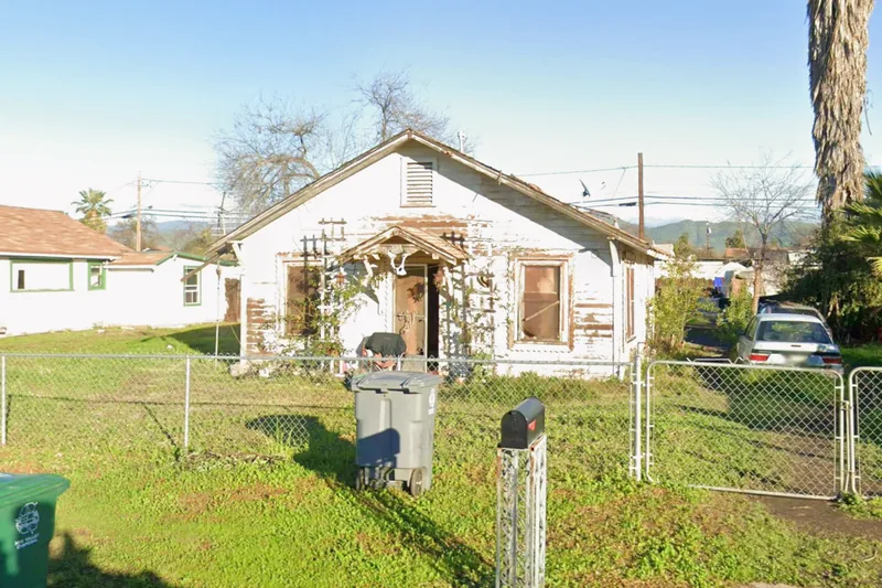 A derelict house on the Fresno County tax-sale list, peeling paint and a boarded window behind a chain-link fence